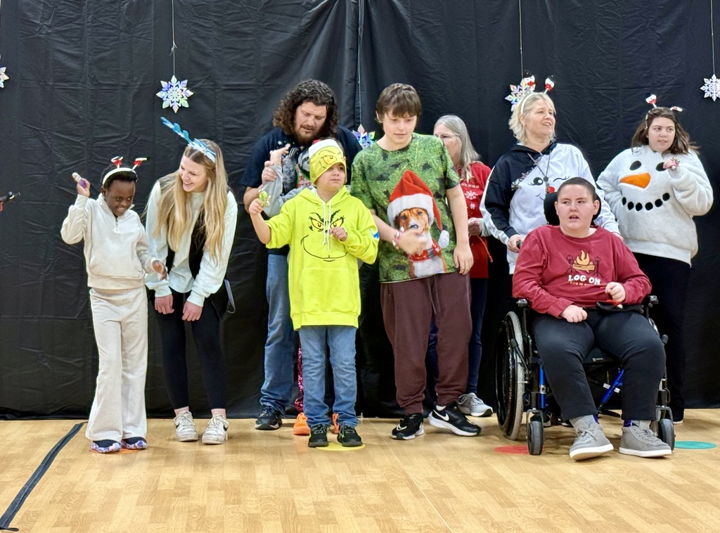 A group of festively dressed staff and students stand in a line. One is in a wheelchair with a staff member behind him. 