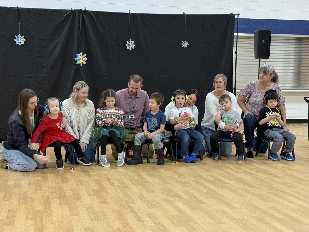 A line of preschool students sit in small chairs with adult staff members behind them. 