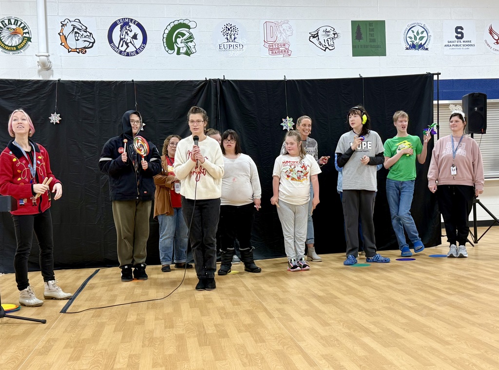  A group of teenage students stand in a line. One of them holds a microphone to talk to the audience. Some of the students have festive holiday clothing  on. 