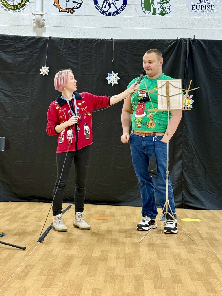 A teacher with a brightly colored Christmas sweater on holds a microphone out for a students dressed in a "Grinch" t-shirt. There is a music stand with papers in front of him. 