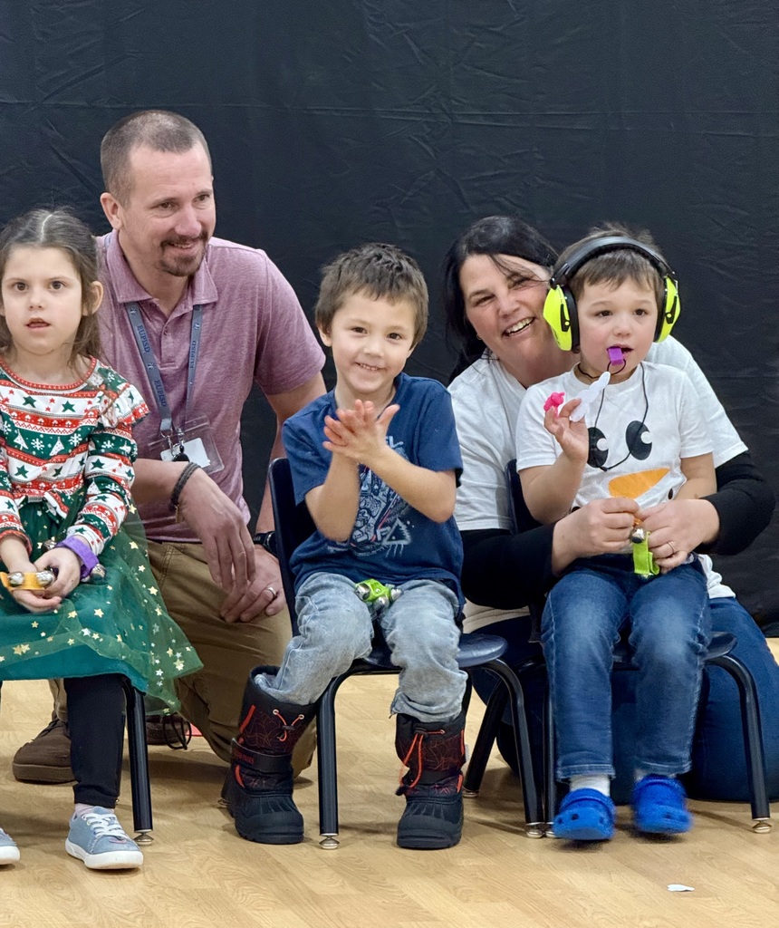 3 preschool children sit in small chairs. One is wearing noise cancelling headphones and there are two staff members  smiling and kneeling behind them. 