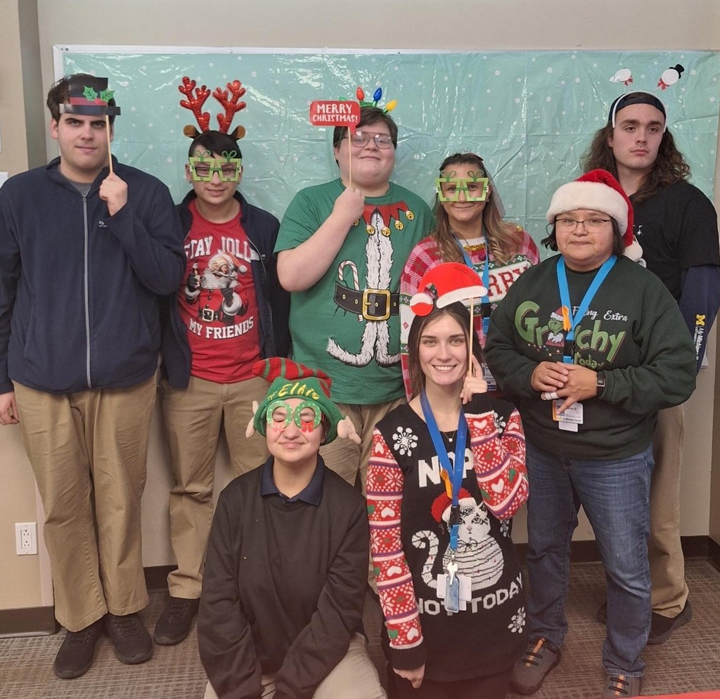 A group picture. Smiling students and staff pose with holiday photo props. They are dressed in festive shirts. Some have fun holiday decor headbands and/or glasses. 