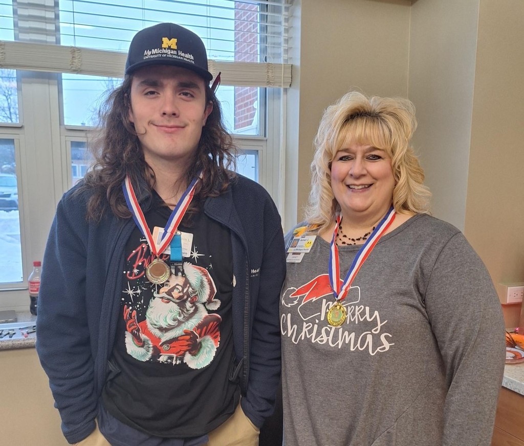 A smiling intern in a Santa shirt smiles with a staff member. Both are wearing medals from winnning the Gingerbread House decorating contest. 