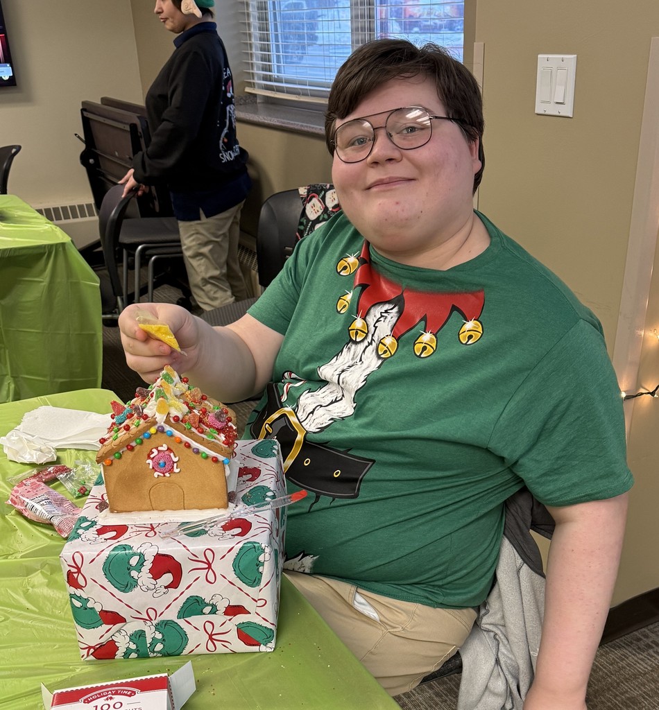 A student in a festive Christmas t-shirt smiles and holds a small tube of frosting. He is decorating a colorful gingerbread house. 