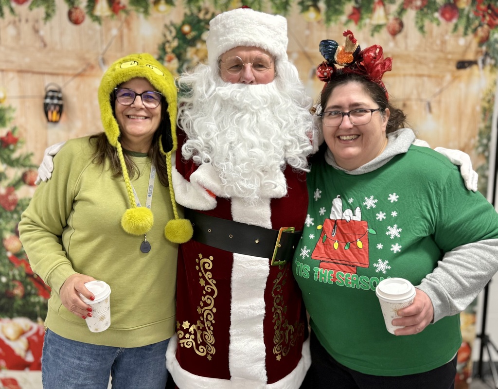 Two staff members in festive holiday clothing pose and smile with Santa Claus. One has a festive headband and  Snoopy shirt and one has a Grinch hat. 