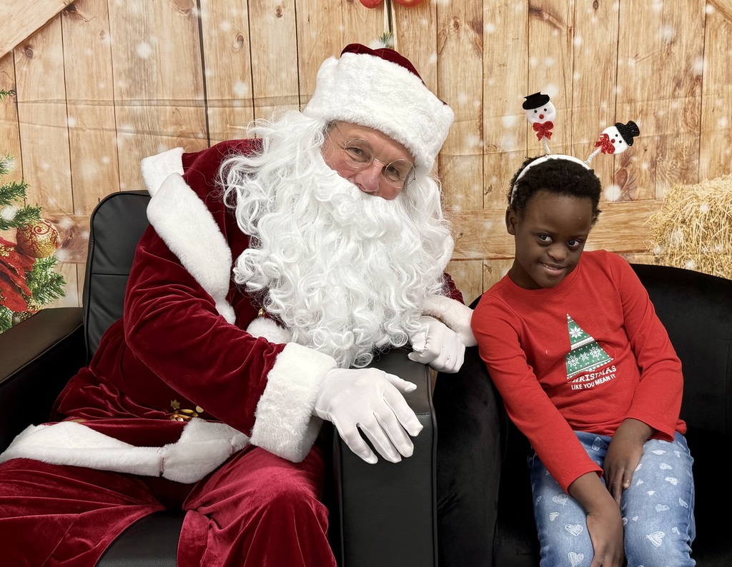 A students sits next to Santa, grinning. Santa leans towards her, smiling. There is a festively decorated barn door background behind them. 
