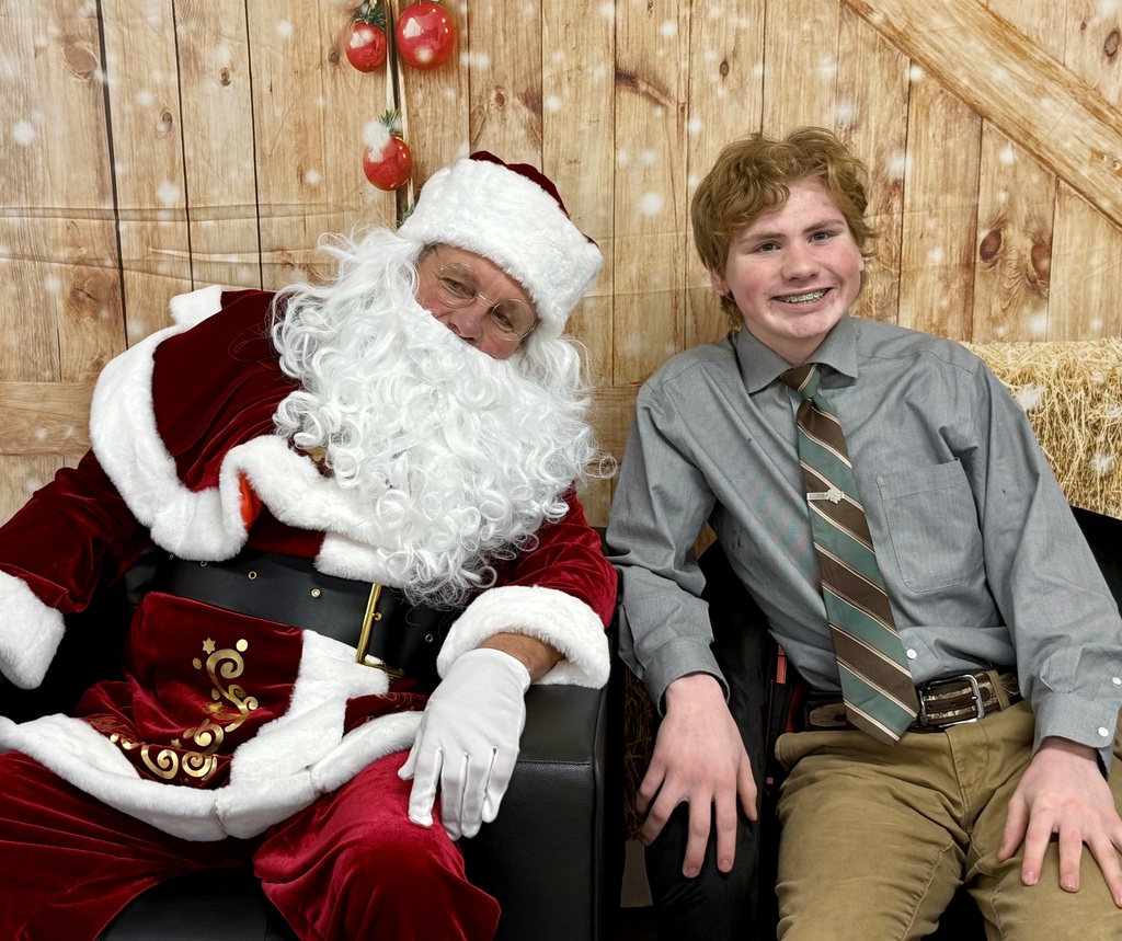 A smiling student in a shirt and tie sits smiling next to Santa. There is a festively decorated barn door background behind them. 