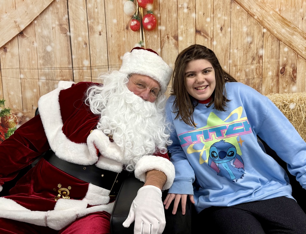 A students sits next to Santa, smiling. She and Santa lean towards each other, posing for the camera. There is a festively decorated barn door background behind them. 