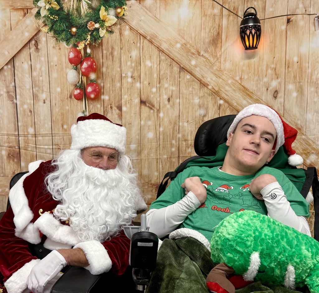 A student in a wheelchair smiles and sits next to Santa. There is a festively decorated barn door background behind them. 