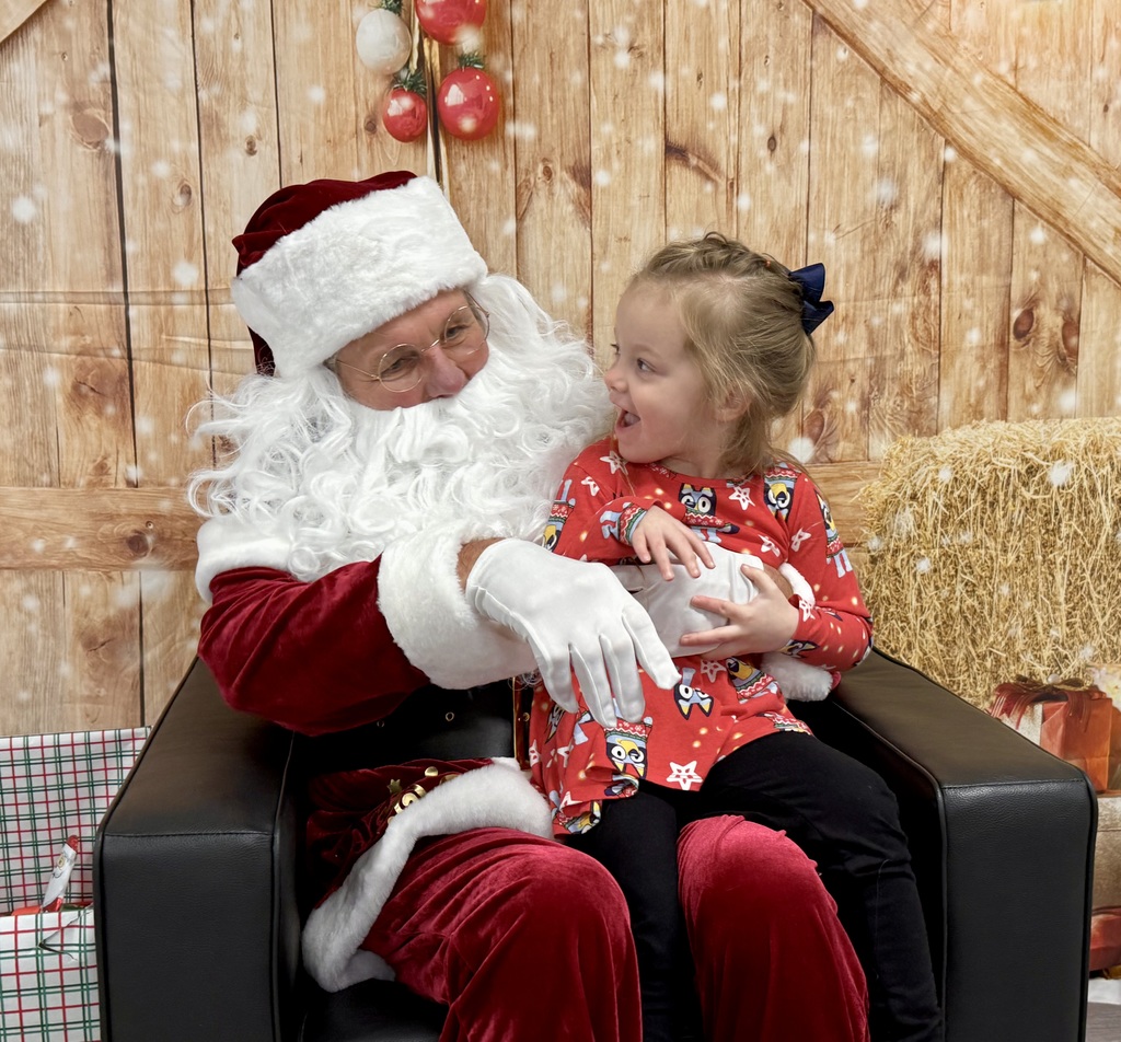 A preschool student sits on Santa's lap and smiles. There is a festively decorated barn door background behind them. 