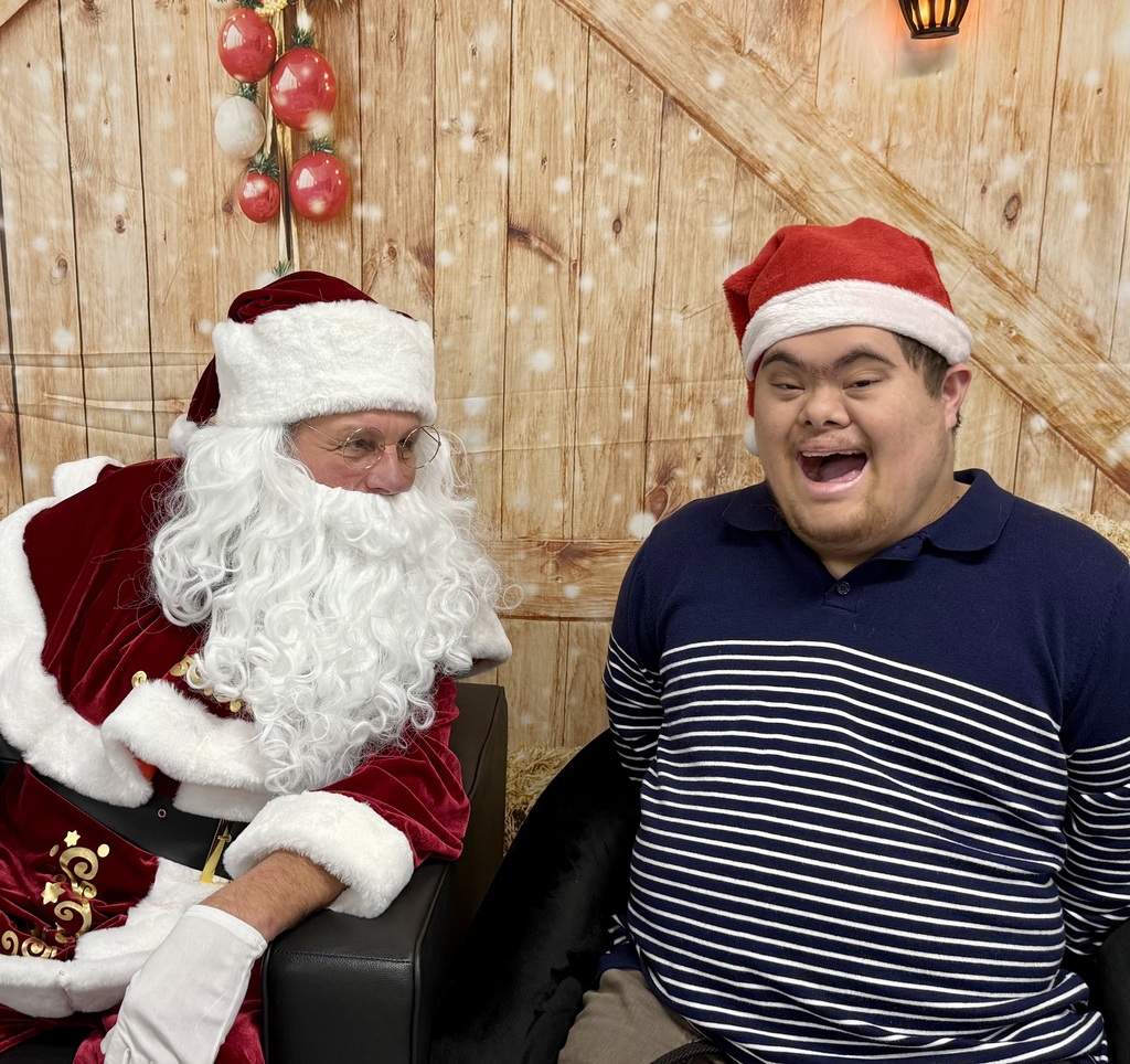 A student smiles so big his mouth is open. He sits next to Santa, who is turned to smile at him.  There is a festively decorated barn door background behind them. 