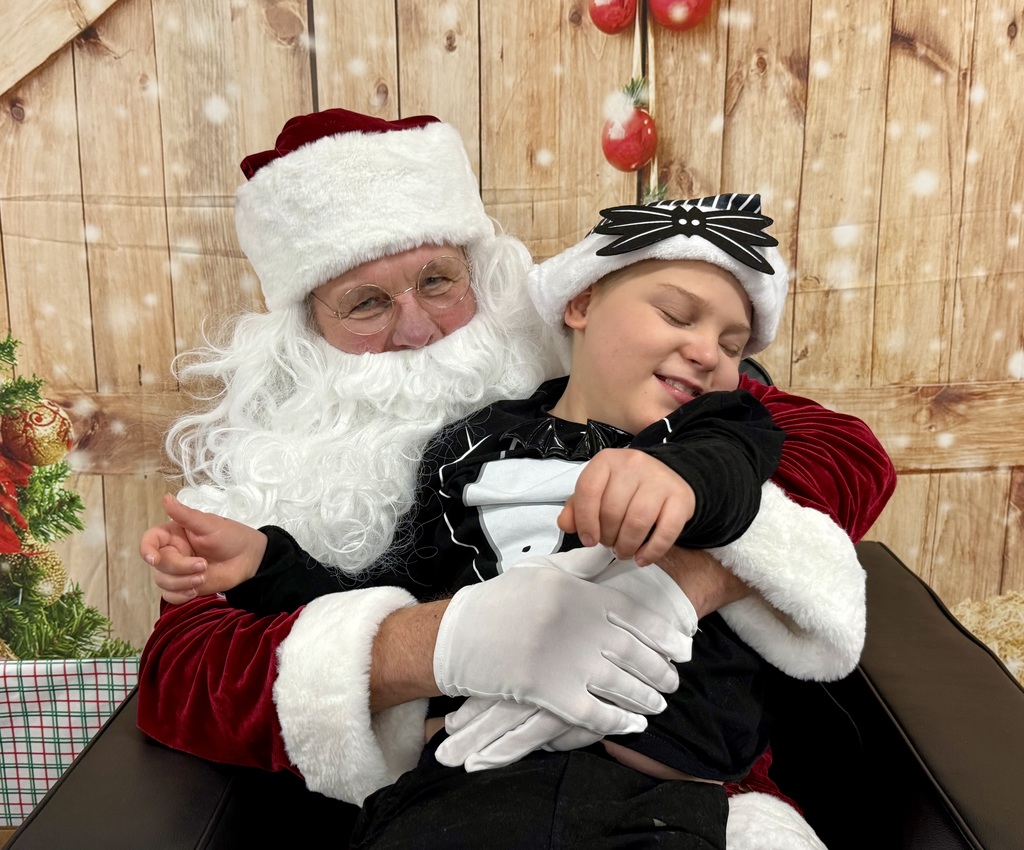 A preschooler sits with Santa. There is a festively decorated barn door background behind them. 