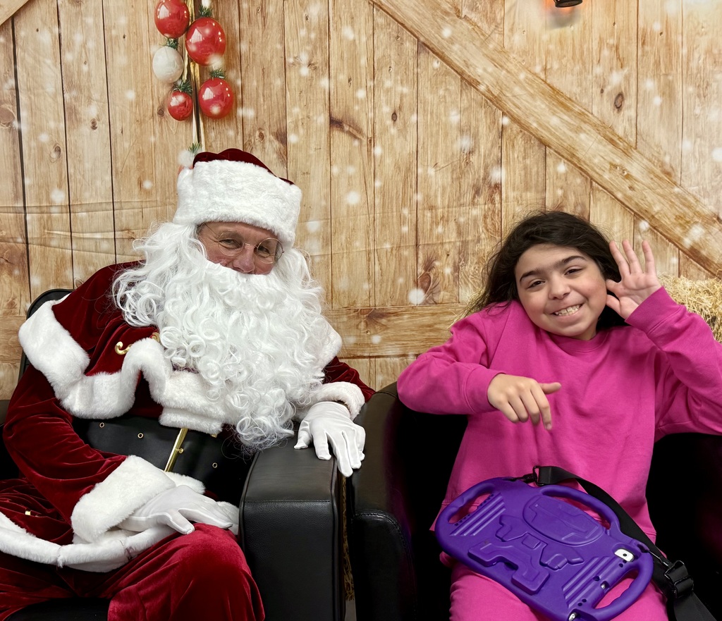 A student grins and waves while sitting in a chair next to Santa. There is a festively decorated barn door background behind them. 