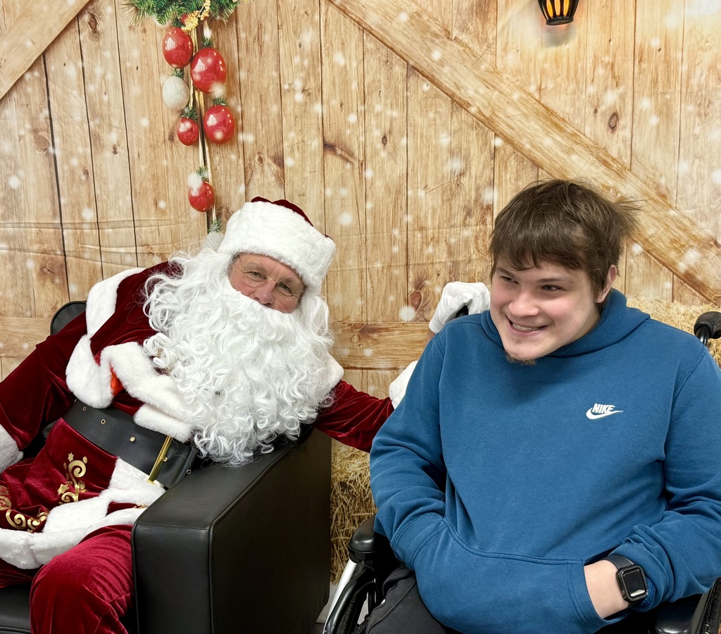 A student in a wheelchair sits next to Santa. Both are smiling at the camera.  There is a festively decorated barn door background behind them. 