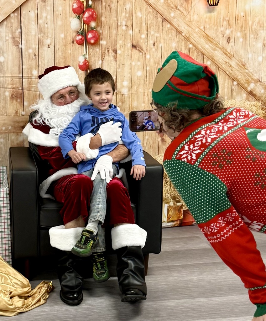 A grinning preschooler sits on Santa's lap. You can view the scene through the staff person who is taking the pictures.  There is a festively decorated barn door background behind them. 