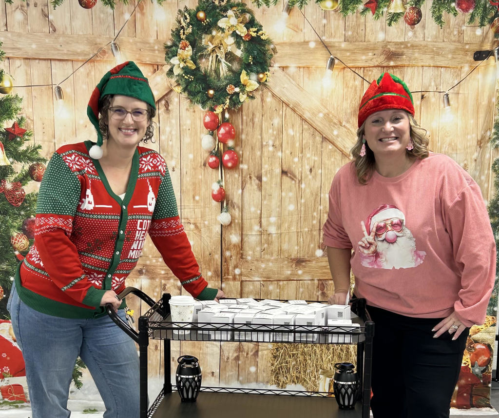 Two staff members pose for a pic. There are wearing festive holiday sweaters and red and green elf hats. There is a festively decorated barn door background behind them. 