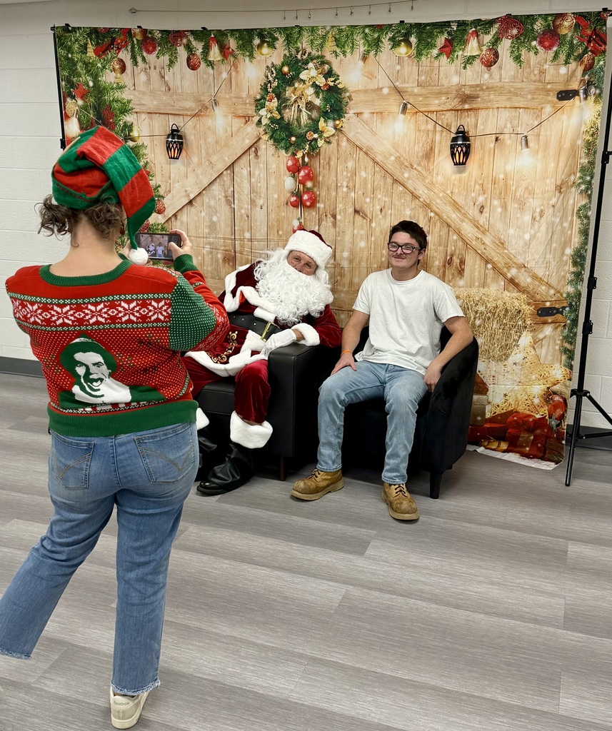 A staff member in an "Elf" sweater takes a picture of a student sitting in a chair next to Santa.There is a festively decorated barn door background behind them. 