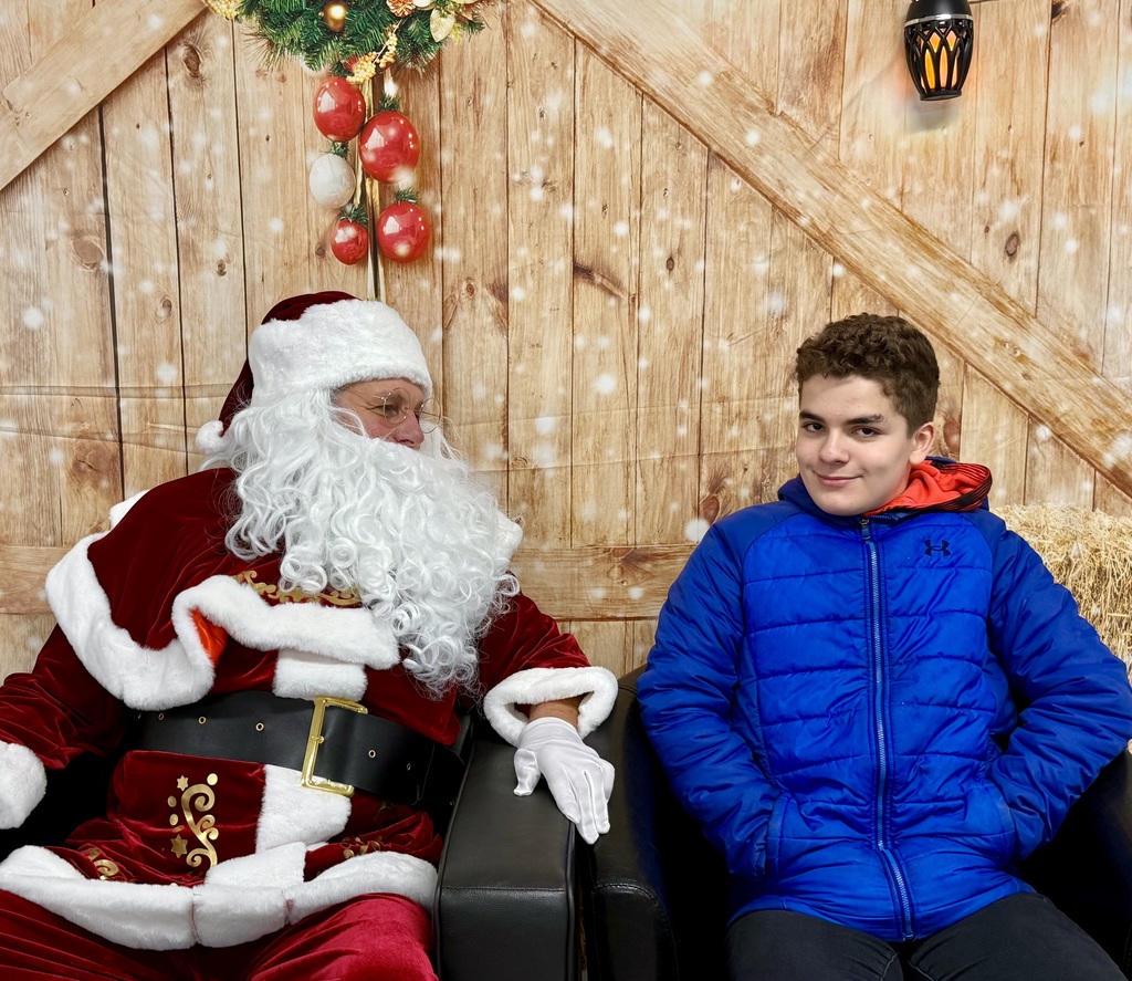 A student looks at the camera. Santa sits next to him and looks over. There is a festively decorated barn door background behind them. 