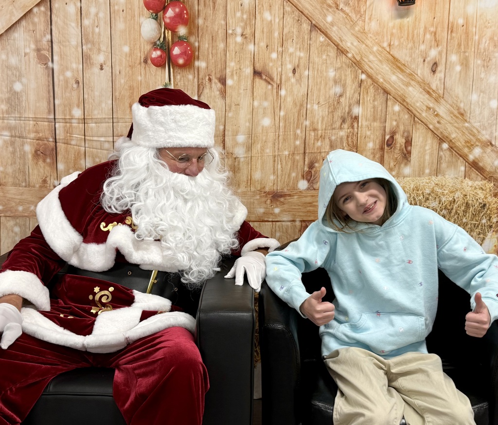 A smiling student sits in a chair next to Santa. She is giving the camera a thumbs up. There is a festively decorated barn door background behind them. 