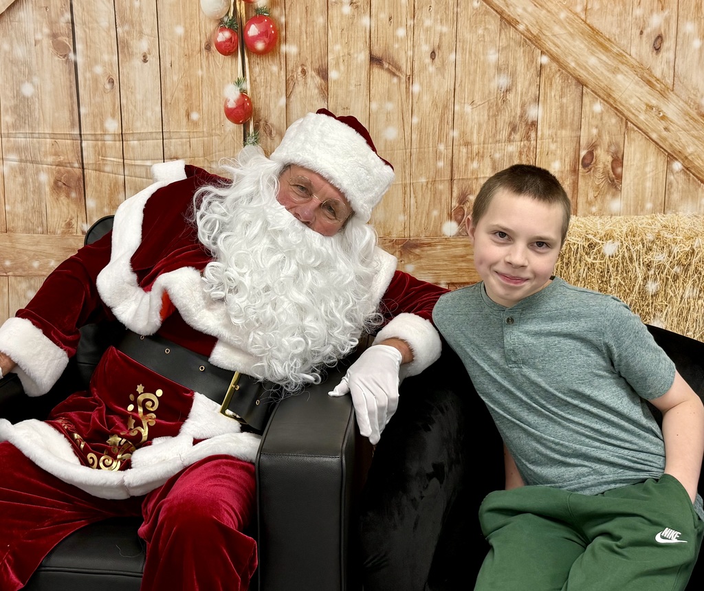 A student smiles as he sits in a chair. They lean towards each other for the camera. There is a festively decorated barn door background behind them. 