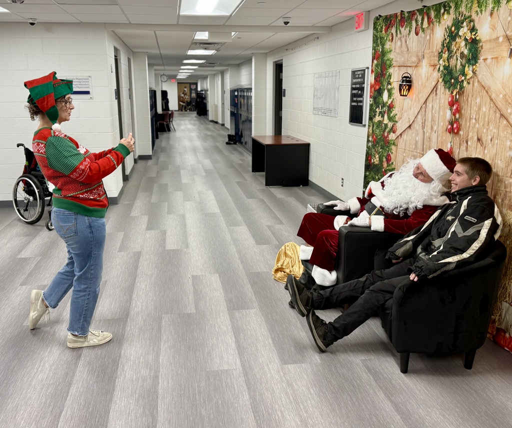 Sideview of a staff person taking a picture of Santa sitting next to a student. Santa and the student lean towards each other for the photo. There is a festively decorated barn door background behind them. 