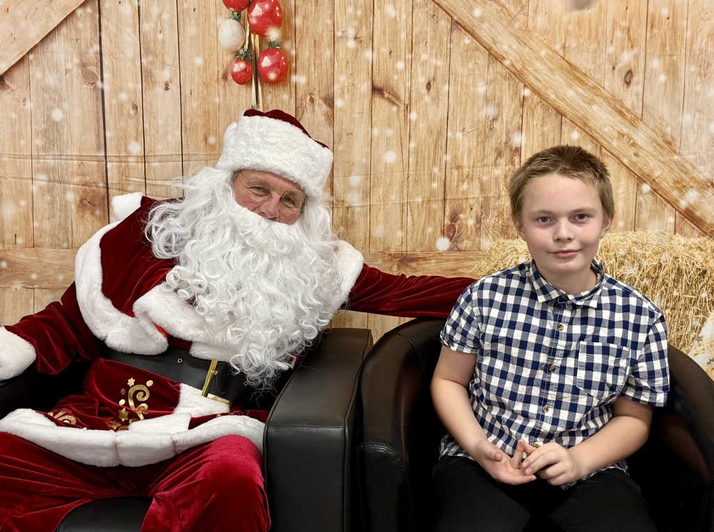 A student sits next to Santa. He is smiling at the camera. There is a festively decorated barn door background behind them. 