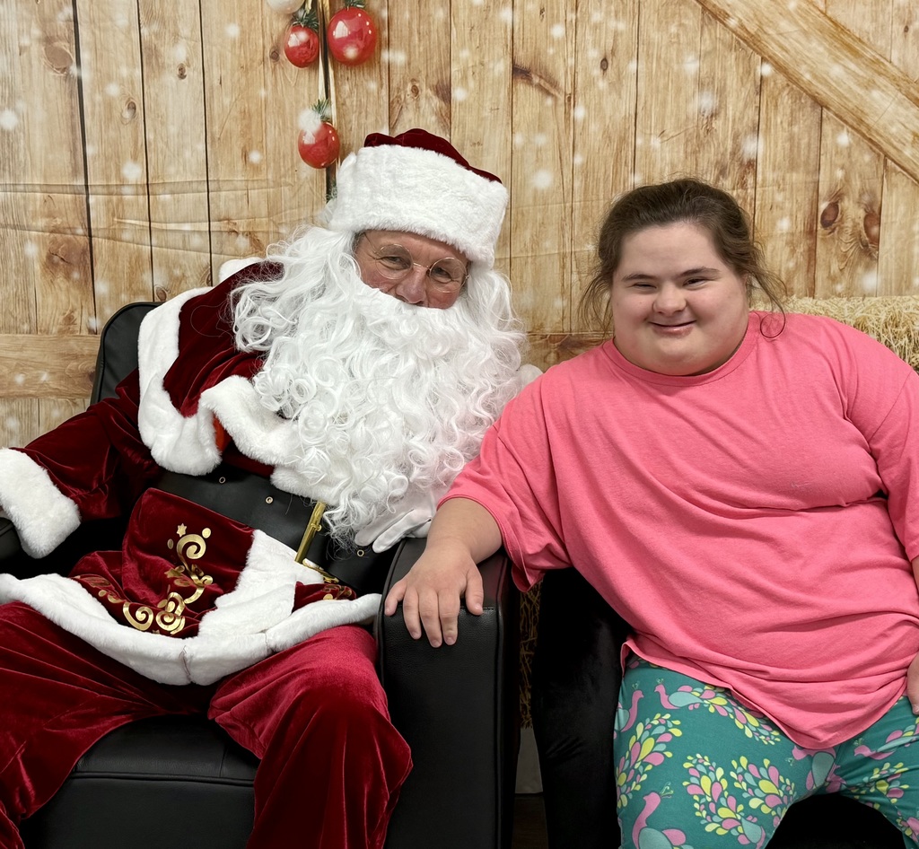 A smiling student in a bright pink shirt smiles as she leans towards a smiling Santa. There is a festively decorated barn door background behind them. 