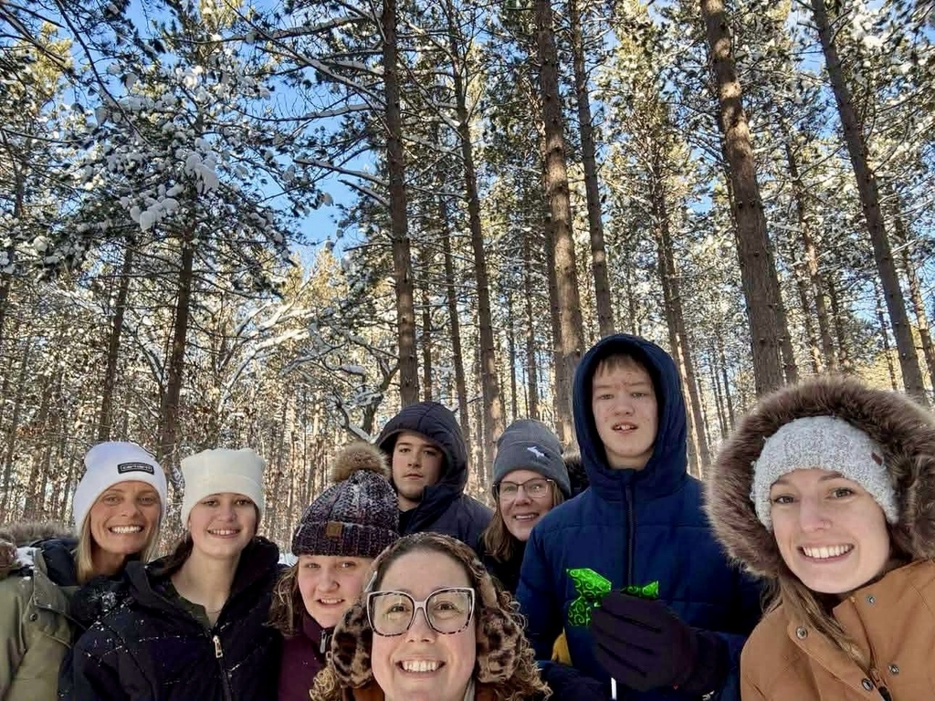 A group of students and staff smile for a group selfie. There is blue sky and tall trees in the woods behind them. 