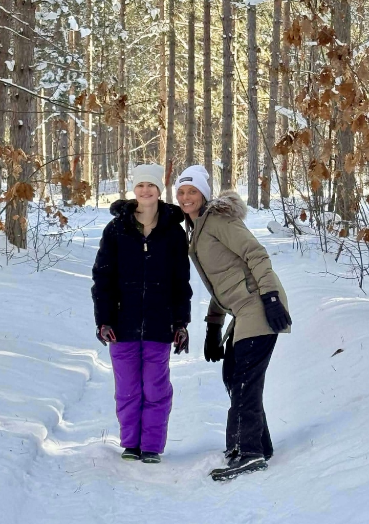 A teacher and her student stand in snow gear in the middle of a snowy trail. They smile for the camera. The sun filters through the tall tress in the woods behind them. 