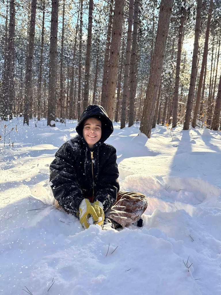 A student in snow gear sits in the snow. He smiles as he scoops up snow between his hands. There is sun filtering through the tall trees in the woods behind him. 