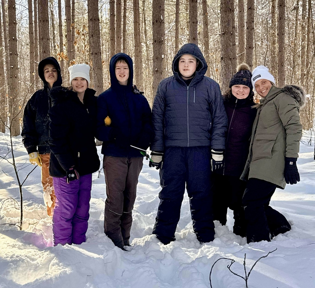 A line of students in snow gear stand in a line in the snow. There is snow filtering through the tall trees in the woods behind them. 
