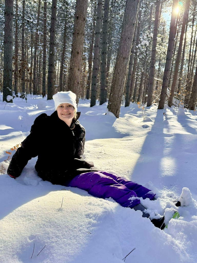 A student in snow gear sits in the snow. She smiles at the camera. 