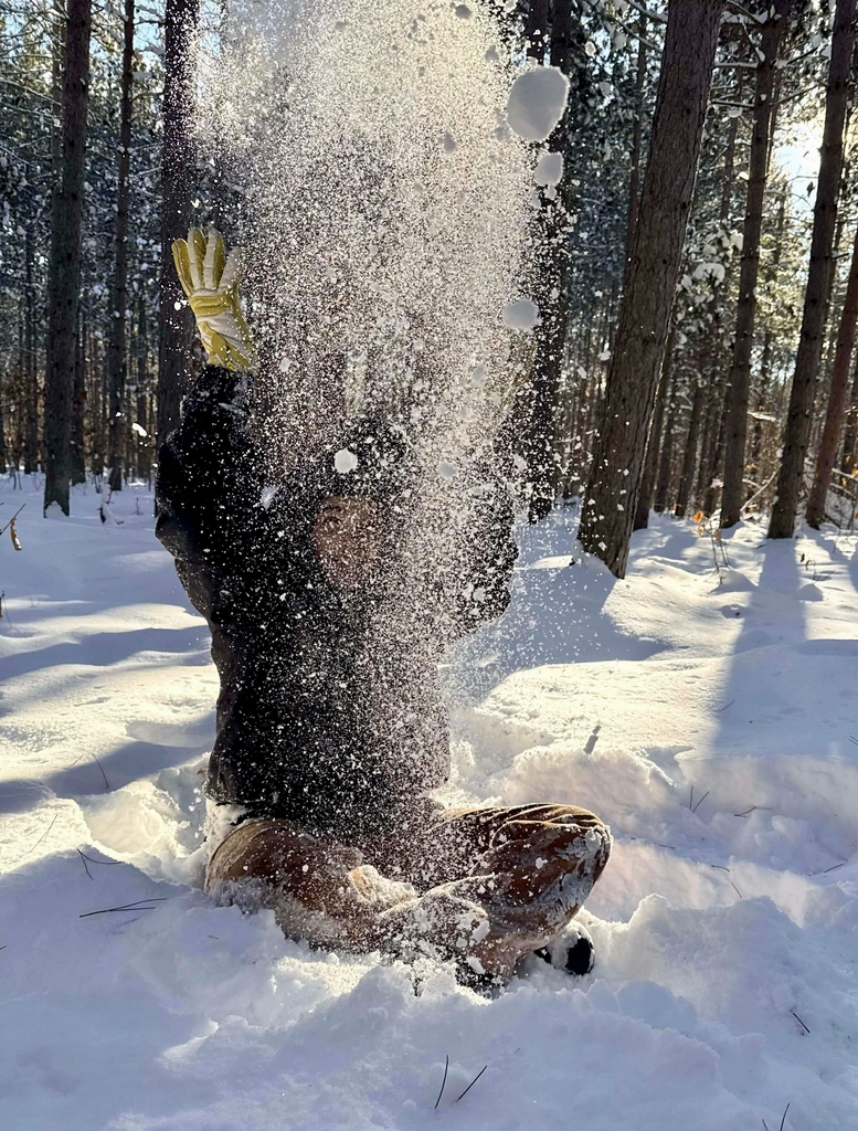 A student in snow gear throws a spray of snow into the air. The sun shines through the trees behind him. 