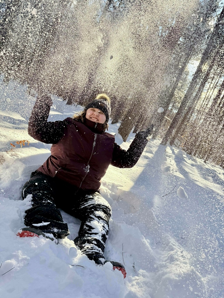 A smiling student in snow gear sits in the snow. She squints as she throws a spray of snow into the air. The sun shines through the trees behind her. 