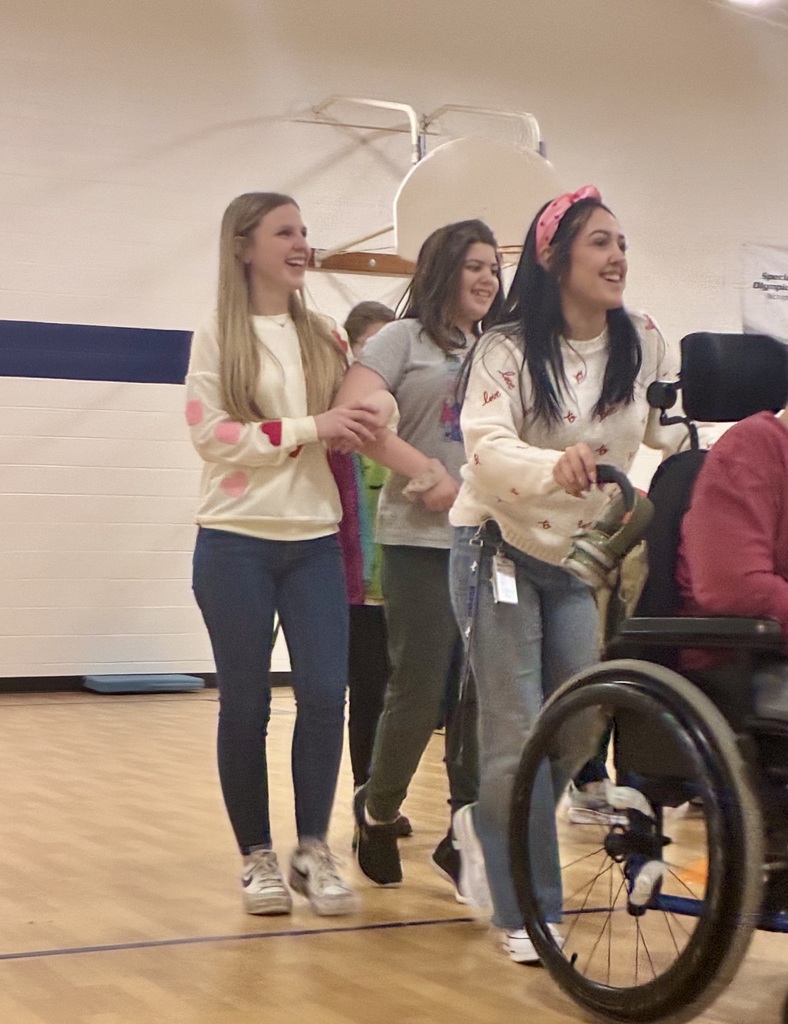 A smiling staff member pushes the wheelchair of a student who is out of frame. Behind her, another smiling staff member with long blond hair and pink and red hearts on her sweater walks arm and arm with a smiling student. They are in a brightly lit school gym. 