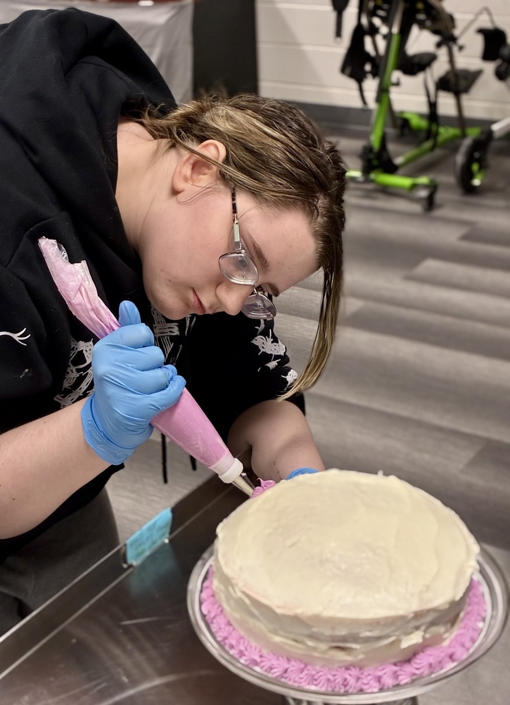 A student bends over a white, frosted cake. She holds a  piping bag filled with pink frosting in her blue gloved hands. She is piping decorations on the edge of the cake. 