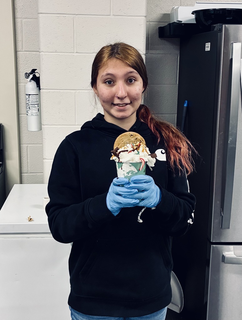 A young woman stands, smiling for the camera. In her blue, food preparation gloves, she is holding a "loaded hot chocolate." A chocolate chip cookie can be seen sticking out of the whipped cream. There is also sprinkles, chocolate and a mini candy cane on top. 