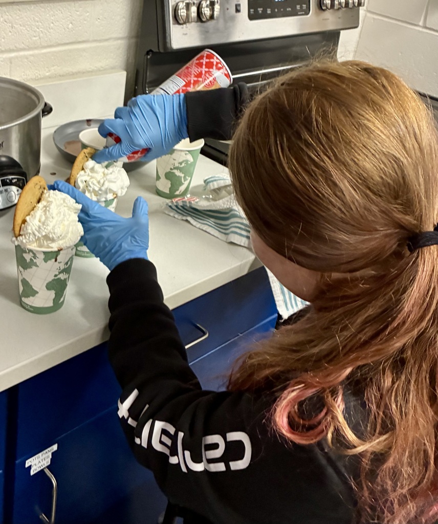 The back of a young woman is facing the camera. She has blue food preparation gloves on and she is spraying whipped cream into two cups. A chocolate chip cooke sticks out of the whipped cream of each cup. 