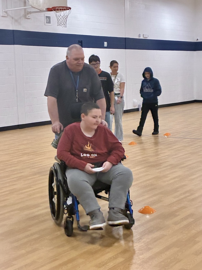A staff person pushes a young man in a wheelchair around a circle of orange cones on the floor. There are students in line behind them. They are in a brightly lit school gym. 