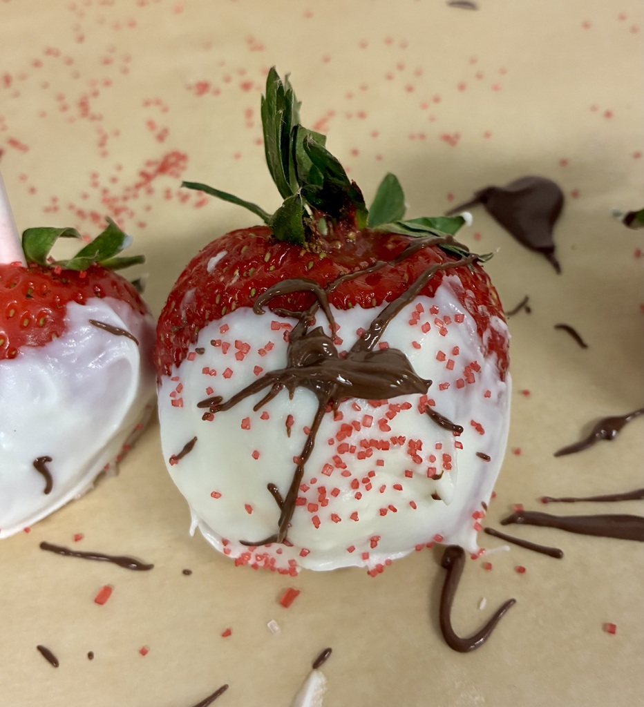 A bright red strawberry is dipped in white chocolate and covered in red sprinkles. There is also brown milk chocolate drizzle on it. Sprinkles and chocolate surround the strawberry on the parchment paper. 