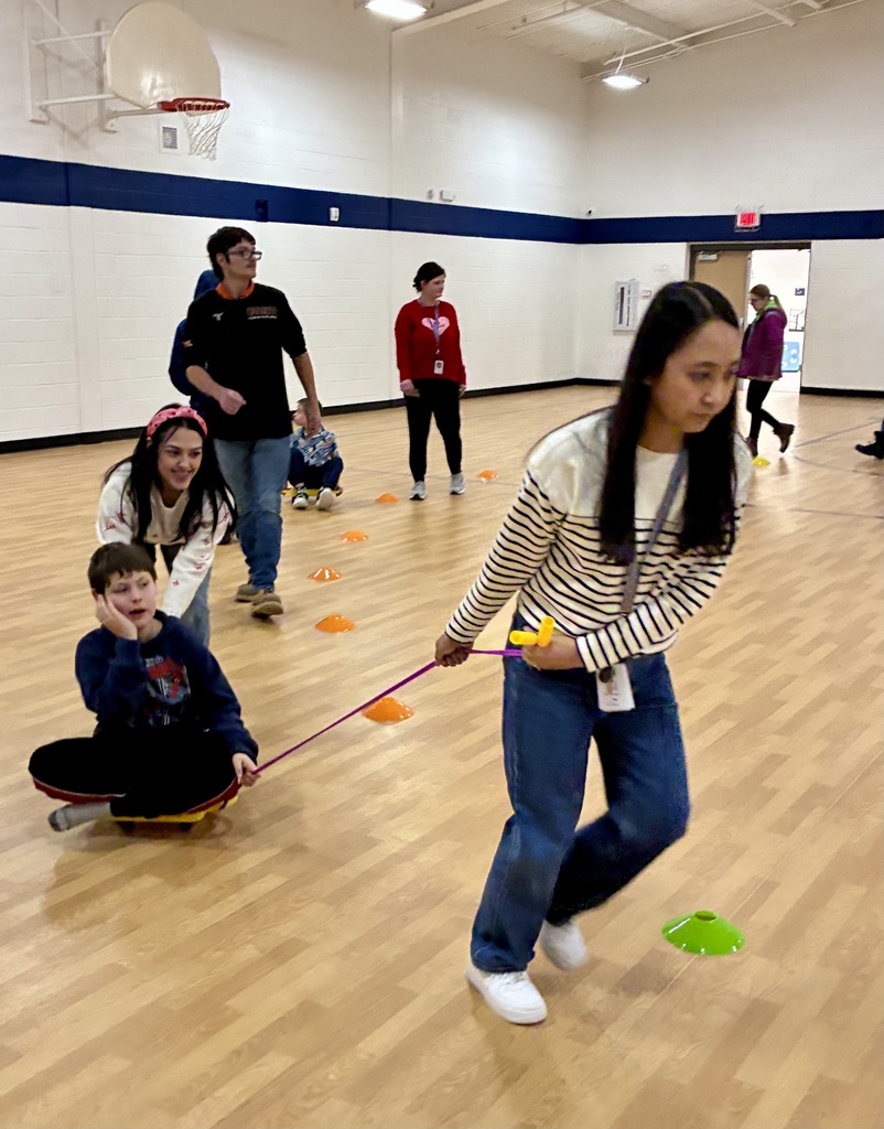 A staff member pulls on a jumprope held by a student on a 4 wheeled scooter. Another staff person, smiling , has her hands out to support his back. There is a brightly lit school gym in the background. 