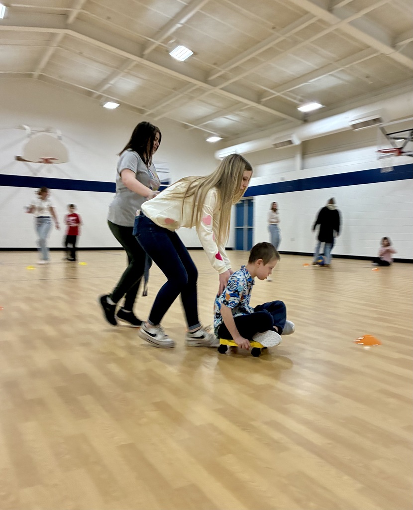 A staff member is bending over and has her hands out to support the back of a student in a brightly colored shirt. She is pushing him forward toward a brightly colored orange cone on the floor of a brightly lit school gym.