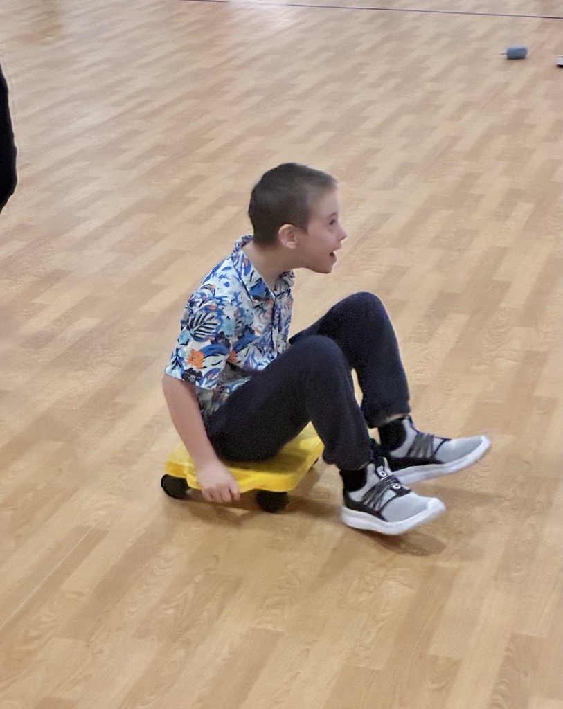 A student laughs, mouth open. He is on a small 4 wheeled scooter on the hard wood floor of a brightly lit school gym. He has his feet on the floor in front of him to propel himself foward on the scooter. 