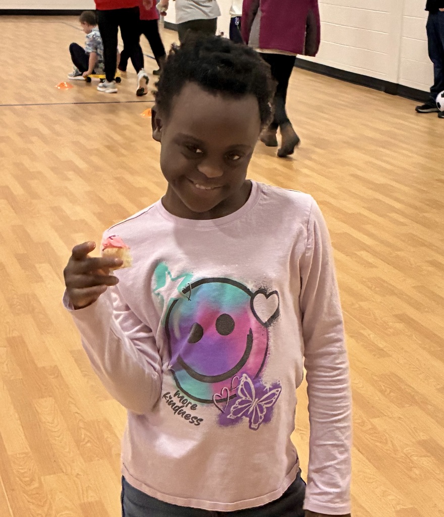 A young, smiley student proudly displays the small cupcake with pink frosting that she won in the cake walk.  She is in a brightly lit school gym.  Her shirt has a big smiley face , butterfly and hearts. It says "more kindness."