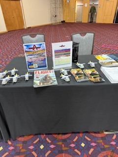 “Recruitment table displaying model airplanes, brochures, and informational signage arranged neatly on a black tablecloth inside a convention-style room.”
