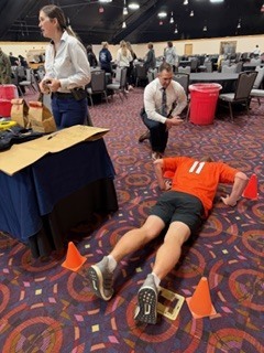 “Person in an orange athletic jersey performing push-ups between orange cones on a patterned carpeted floor inside a large event hall, while two nearby adults observe and tables with supplies are visible in the background.”
