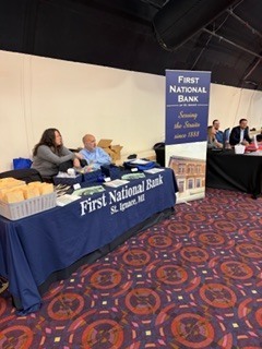 “First National Bank of St. Ignace booth with a blue tablecloth, paperwork, and small boxes, with a tall vertical banner displaying the bank name and location inside an event hall.”