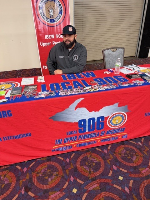 “Close-up view of a union information booth with a red tablecloth reading ‘IBEW Local 906’ and ‘Upper Peninsula of Michigan,’ featuring brochures, stickers, and a standing banner behind the table.”