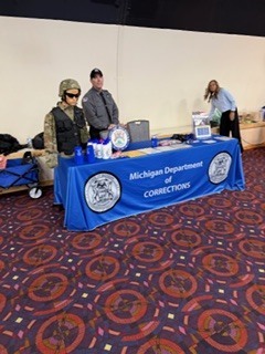 “Michigan Department of Corrections display table with a blue tablecloth, informational materials, and demonstration equipment, set up inside a large indoor event space.”