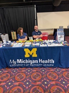 “Information booth with a blue tablecloth labeled ‘My Michigan Health – University of Michigan Health,’ displaying pamphlets, giveaway items, and signage, with two adults seated behind the table.”